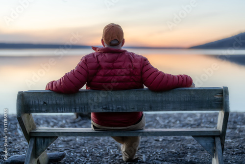 Winter scene of a man with red jacket on a bench looking out over a lake at sunset. Taking time for personal reflection, introspection, thinking about the past or the future. 
