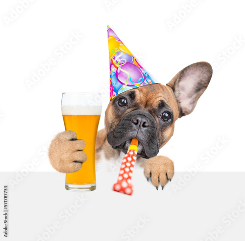 Fototapeta Naklejka Na Ścianę i Meble -  French bulldog puppy wearing party cap blows into party horn, looks above empty white banner and holds glass of beer. isolated on white background