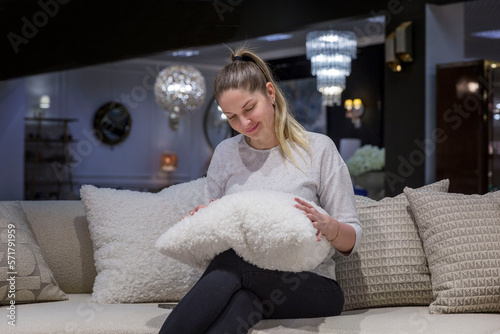 Foto Smiling young woman is sitting on a sofa in a furniture store with a fluffy decorative pillow made of astrakhan in her hands