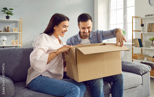 Smiling happy young family is sitting together on sofa at home in cozy living room and unpacking large cardboard box. Harmony in relationships, sincere feelings, joy, positivity, satisfaction, fun.