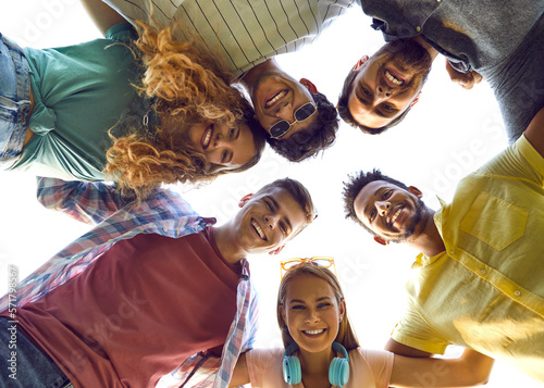 Group of diverse friends have fun together. Six different beautiful young mixed race people standing in circle, hugging and looking down with happy smiling face expressions. Low angle, view from below