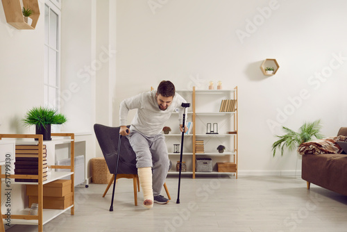 Fototapeta Young man with a broken leg in a plaster cast tries to stand up off his chair and walk with a stick and crutches in the living room at home