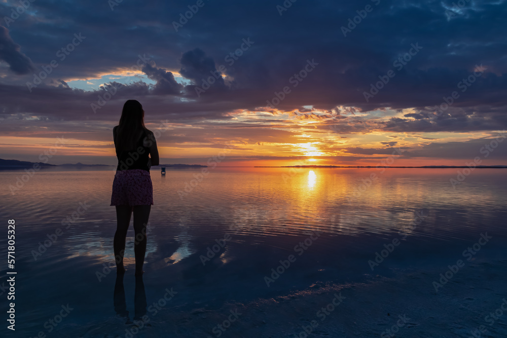 Silhouette of woman walking into the sunrise of lake Bonneville Salt Flats, Wendover, Western Utah, USA, America. Dreamy red colored clouds mirroring on the water surface creating romantic atmosphere