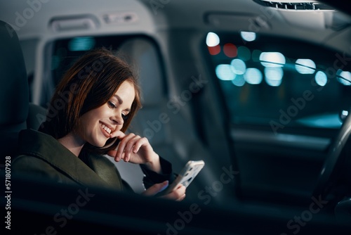 a close horizontal portrait of a stylish, luxurious woman in a leather coat sitting in a black car at night in the passenger seat, thoughtfully looking at her smartphone with her hand near her face © SHOTPRIME STUDIO