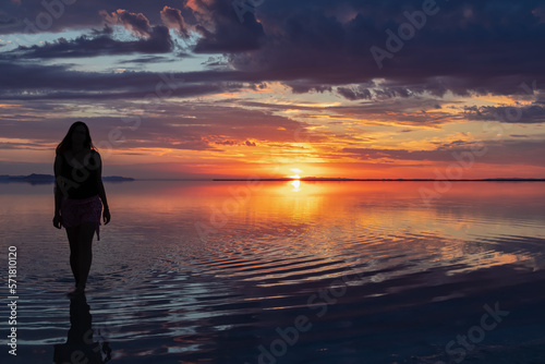 Silhouette of woman walking into the sunrise of lake Bonneville Salt Flats, Wendover, Western Utah, USA, America. Dreamy red colored clouds mirroring on the water surface creating romantic atmosphere © Chris