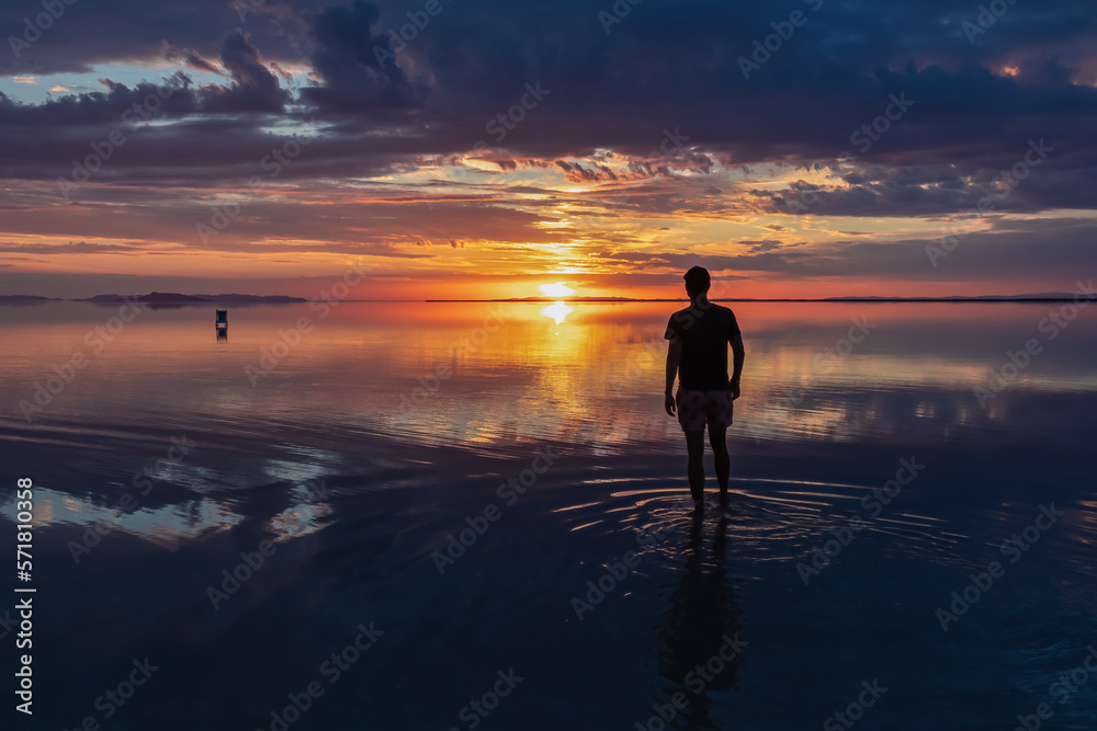Silhouette of man walking into the sunrise of lake Bonneville Salt Flats, Wendover, Western Utah, USA, America. Dreamy red colored clouds mirroring on the water surface creating romantic atmosphere