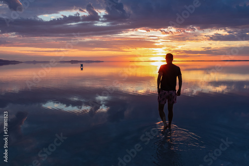 Silhouette of man walking into the sunrise of lake Bonneville Salt Flats, Wendover, Western Utah, USA, America. Dreamy red colored clouds mirroring on the water surface creating romantic atmosphere