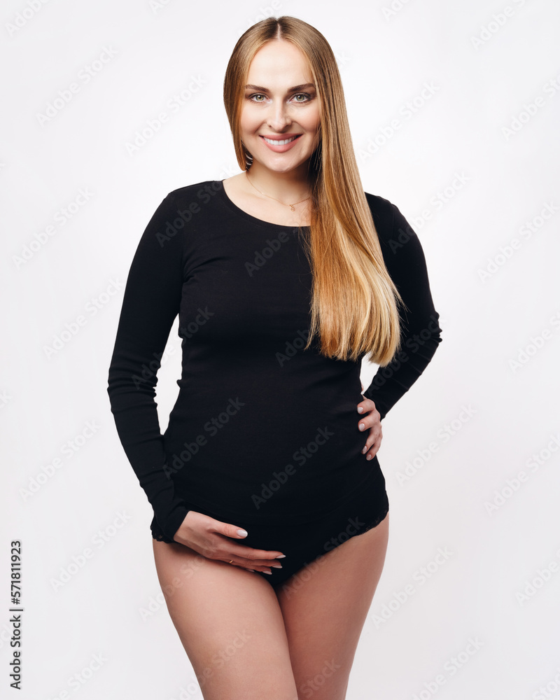 Smiling happy pregnant young woman, girl in black tight-fitting bodysuit isolated on light background. Studio picture. Smiling happy pregnant young woman, girl in black tight-fitting bodysuit isolated on light background. Studio picture.
