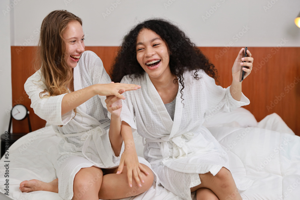 Young happy diversity ethnic beautiful woman in bathrobe sit on bed and playing social media on mobile smart phone and having fun together. Women lesbian couple daily LGBTQ lifestyle and love concept. Young happy diversity ethnic beautiful woman in bathrobe sit on bed and playing social media on mobile smart phone and having fun together. Women lesbian couple daily LGBTQ lifestyle and love concept.