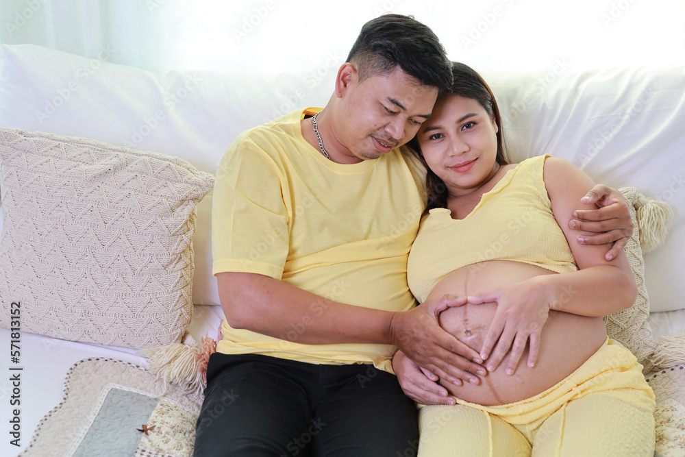 Happy asian adult couple with husband sitting and resting on sofa in living room while holding baby inside a pregnant and smile. Expectant mother preparing and waiting for baby birth during pregnancy. Happy asian adult couple with husband sitting and resting on sofa in living room while holding baby inside a pregnant and smile. Expectant mother preparing and waiting for baby birth during pregnancy.