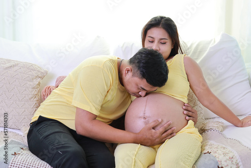 Happy asian adult couple with husband sitting and resting on sofa in living room while kissing a pregnant and smile. Expectant mother preparing and waiting for baby birth during pregnancy.