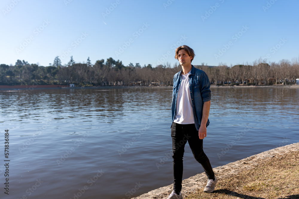 Full body of young male in casual outfit strolling near rippling river on sunny day with cloudless blue sky during free time
