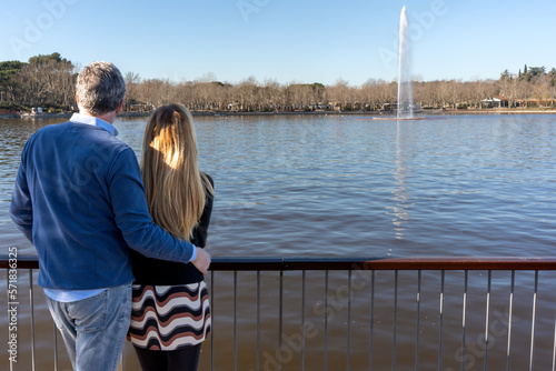 Back view of mature man hugging wife while standing on embankment against lake with fountain in sunny weather during date