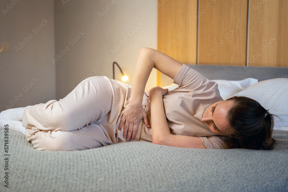 A young woman with a painful expression, holding her stomach, suffering from menstrual pains, lying on a home bed with stomach colic A young woman with a painful expression, holding her stomach, suffering from menstrual pains, lying on a home bed with stomach colic