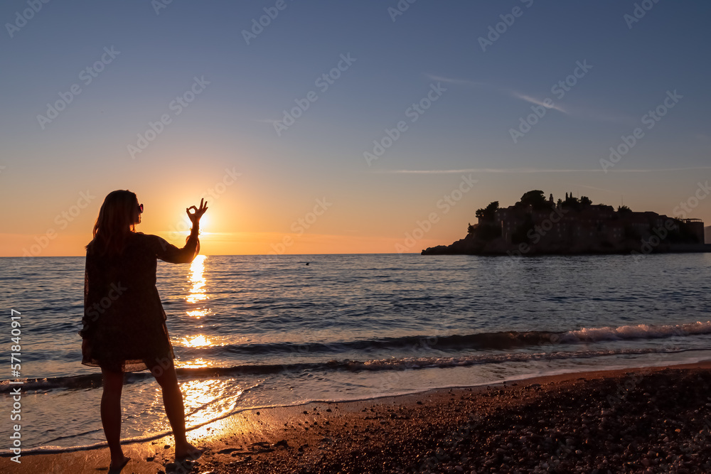Silhouette of barefoot woman on sand beach with scenic view on idyllic island Sveti Stefan at romantic sunset, Budva Riviera, Adriatic Mediterranean Sea, Montenegro, Europe. Summer vacation at seaside