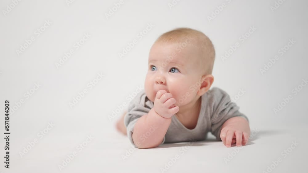 Portrait of a baby. A baby lying on his stomach looking into the camera licking his fingers, shot in close-up against a white background, looking at the cameraman Portrait of a baby. A baby lying on his stomach looking into the camera licking his fingers, shot in close-up against a white background, looking at the cameraman