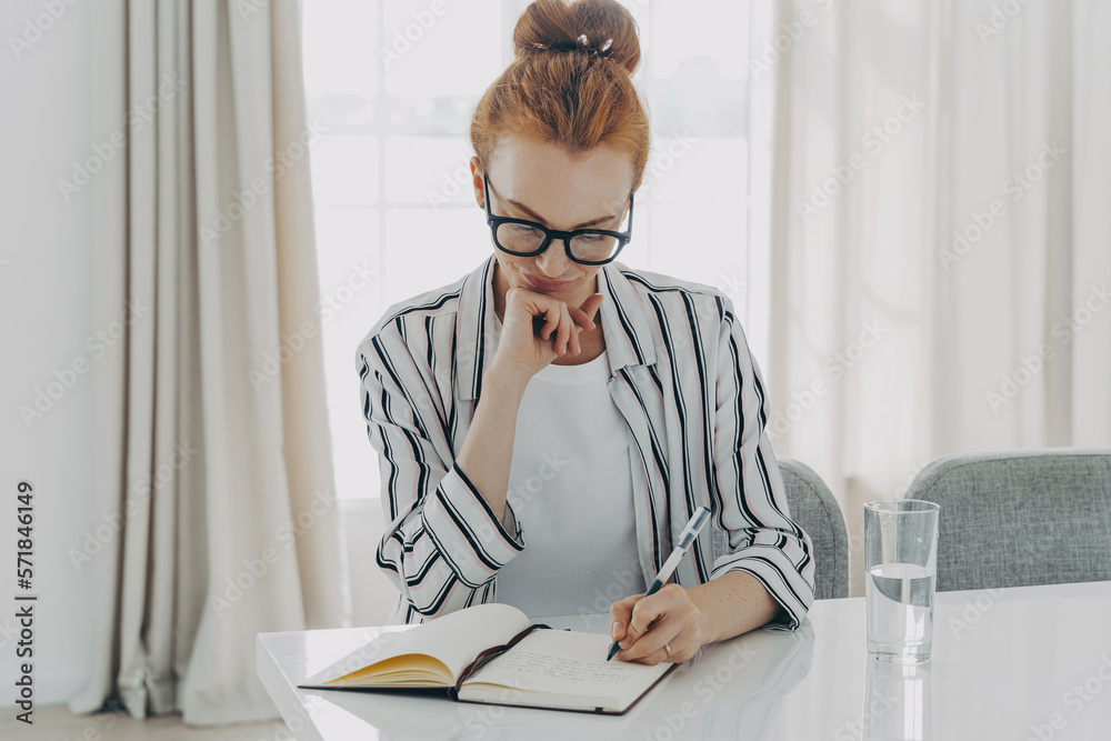 Fototapeta premium Redhead woman in striped shirt takes notes writes down information in notebook