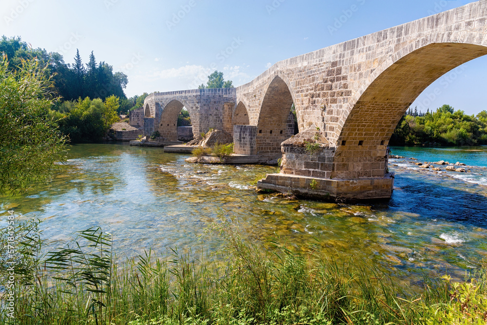 Fototapeta premium Seljuk Bridge in Aspendos. The Eurymedon Bridge. Crooked bridge, reflected in calm water. An ancient building across the Kopruchay river. Antalya region, Turkey.