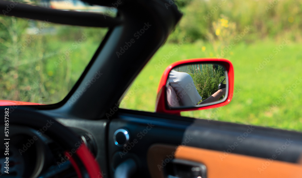 Reflection of woman in white dress near red cabriolet in car mirror close up with selective focus. Road trip enjoying freedom concept 