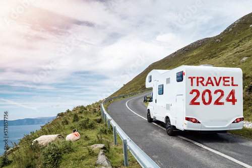 White motorhome with sign travel 2024 moving on a small mountain road and beautiful country side with ocean in foreground. Low cloudy sky. Travel with camper concept. Trip to nature.