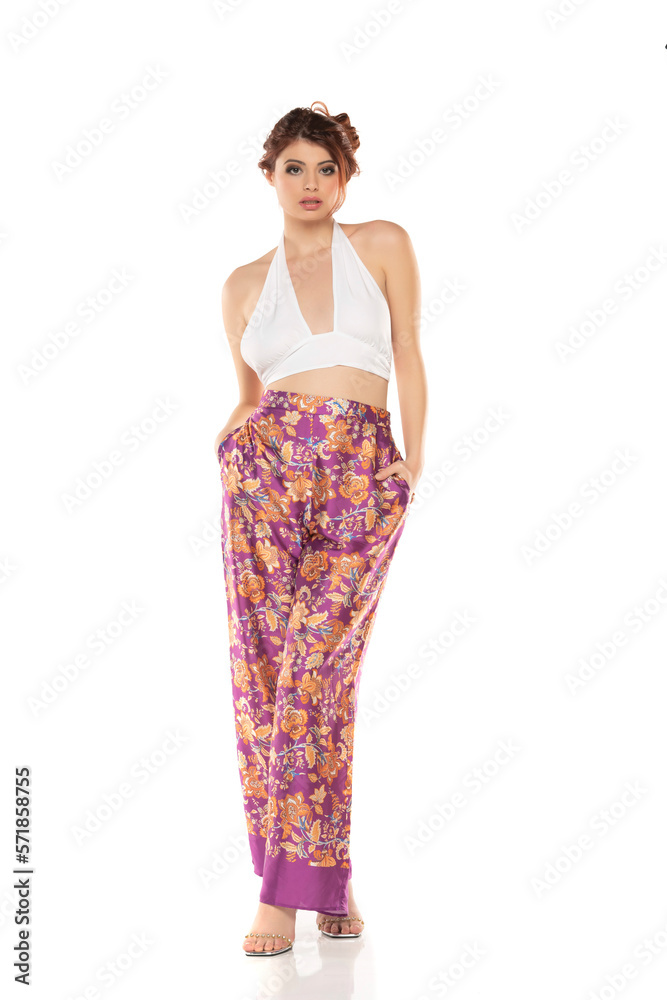 Fashion style studio portrait of beautiful young woman. Floral pattern wide leg pants and white sleevles shirt. Model standing and posing against white background Fashion style studio portrait of beautiful young woman. Floral pattern wide leg pants and white sleevles shirt. Model standing and posing against white background