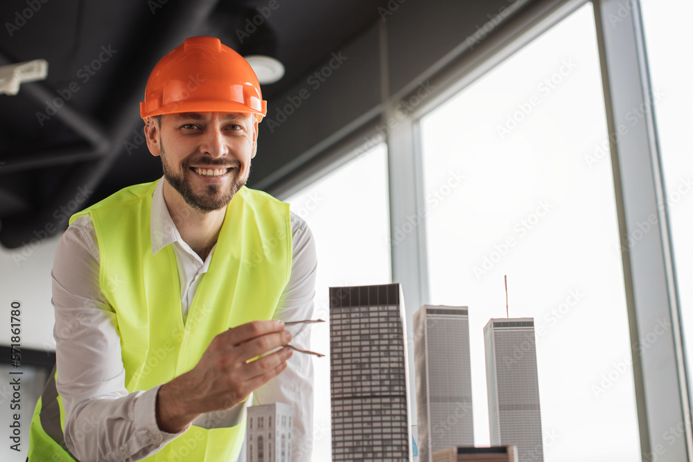 Close up of bearded engineer in protective helmet working on building complex prototype project of residential or business district, measuring city model using compass for drawing at office. Close up of bearded engineer in protective helmet working on building complex prototype project of residential or business district, measuring city model using compass for drawing at office.