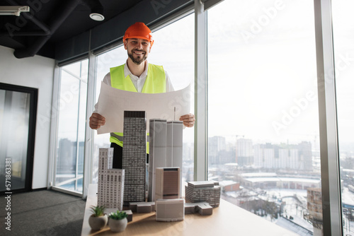 Wallpaper Mural Young builder in hard hat holding blueprint working on building complex prototype project of residential or business district, using city model at open space office with panoramic windows. Torontodigital.ca