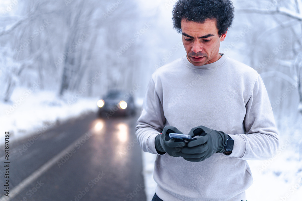 Man looking at his phone and sending text massages, car in the distance approaches there's snow covered scenery in the background Man looking at his phone and sending text massages, car in the distance approaches there's snow covered scenery in the background