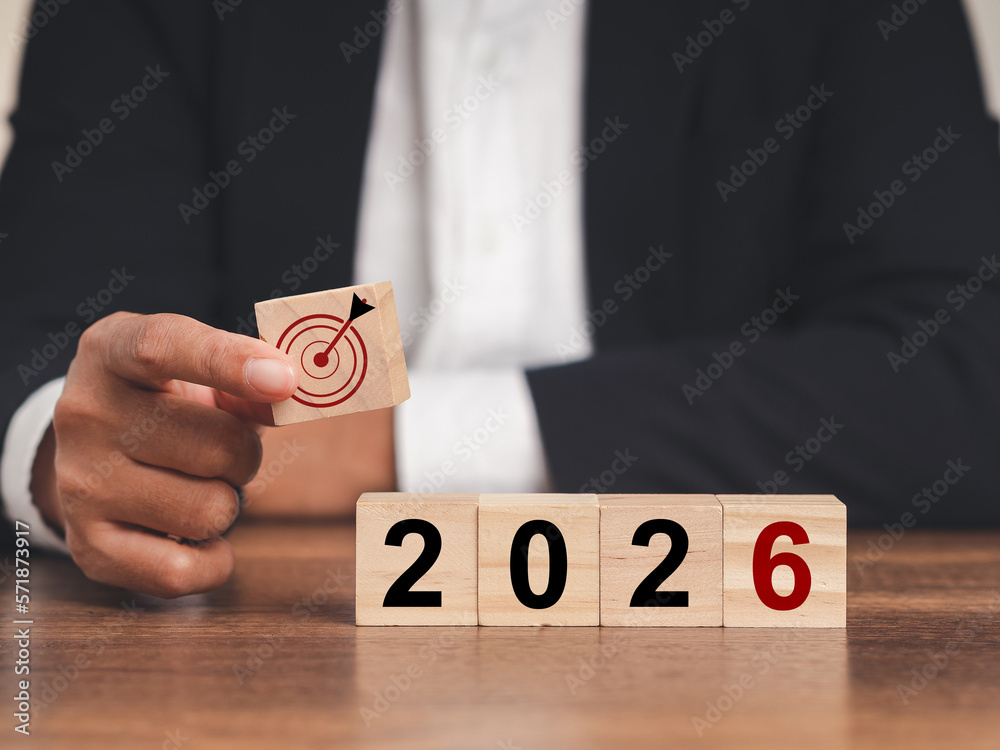 Business planning and strategy in 2026. Hand businessman holding wooden cubes with dartboard icon and wooden cubes with the letters 2026 on a table - obrazy, fototapety, plakaty 