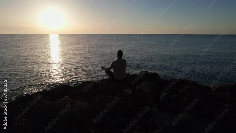 Focused man practicing meditation on a rocky beach summer morning. Yoga man sitting in lotus position near ocean waves. Attractive sportsman doing relaxing asana outdoors. Self care concept. Focused man practicing meditation on a rocky beach summer morning. Yoga man sitting in lotus position near ocean waves. Attractive sportsman doing relaxing asana outdoors. Self care concept.