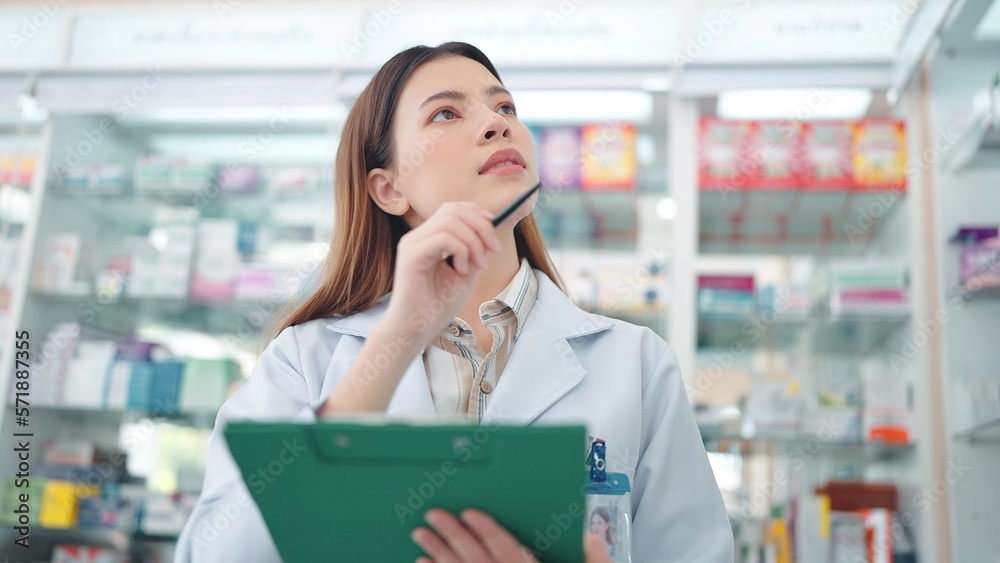 Professional Asian woman pharmacist rechecks products in pharmacy and using noteboard to write information