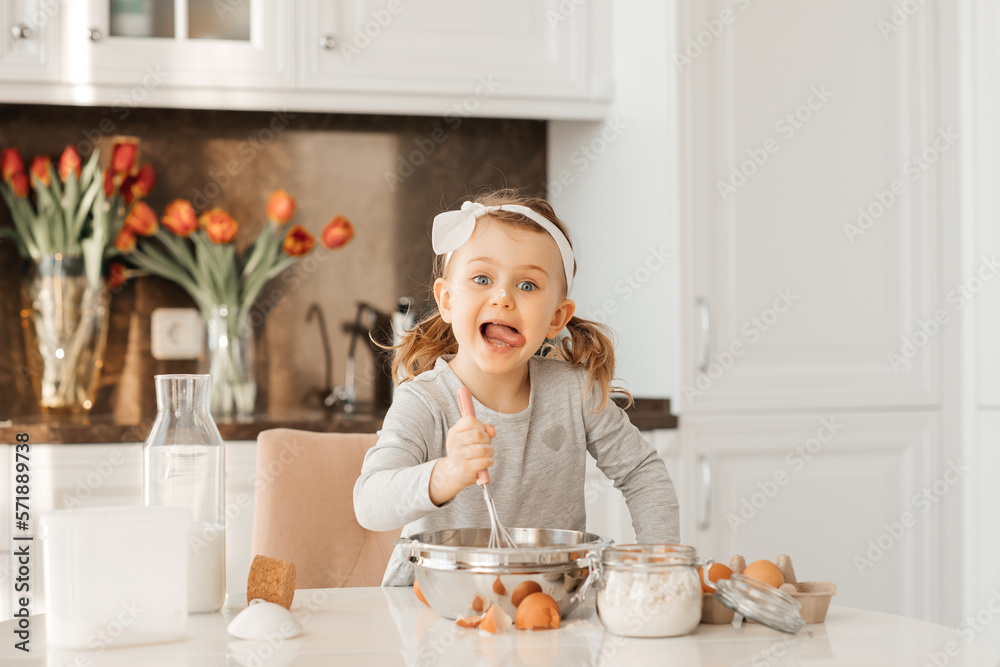 happy excited kid girl baking cake for Easter emotional child cook dessert present for mother day in white sunny kitchen Home bakery. Bottle of milk, eggs, flour are on the table happy excited kid girl baking cake for Easter emotional child cook dessert present for mother day in white sunny kitchen Home bakery. Bottle of milk, eggs, flour are on the table