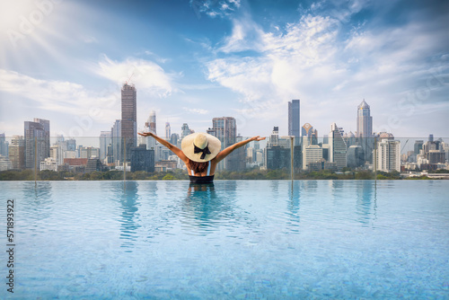 A happy tourist woman with hat enjoys the view over the urban skyline of Bangkok, Thailand, from a swimming pool photo