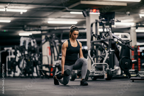 A muscular sportswoman is lifting dumbbells and doing lunges in a gym.