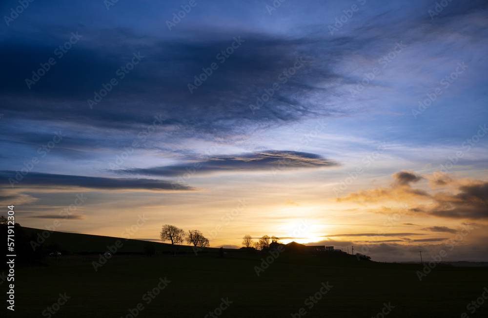 The silhouette of a farm and trees in the English countryside at sunset with dramatic clouds high above in winter, UK.