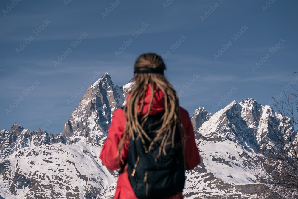 Silhouette of young woman out of focus with long dreadlocks and backpack against background of winter mountain peaks covered with snow