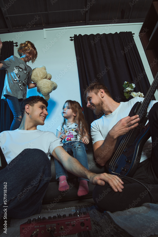 Staged photo. Homosexual couple and their children at home.   The whole family gathered on the couch.  Home concert with a guitar. One of the girls sings with her toy bear. You're doing great, baby!