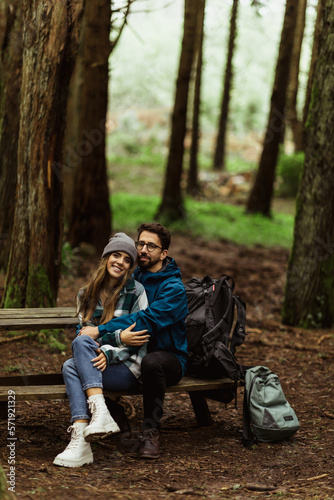 Glad millennial caucasian husband in jacket hugging wife, resting on bench in cold forest with backpack