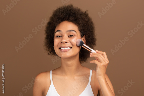 Smiling young black curly lady with perfect skin applies powder or blush with brush on face