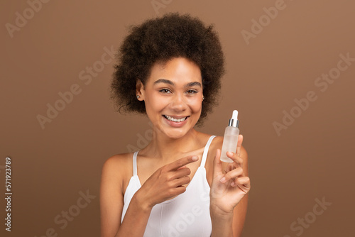 Smiling young black curly lady with perfect skin point finger at jar of oil, serum, recommend moisturizing