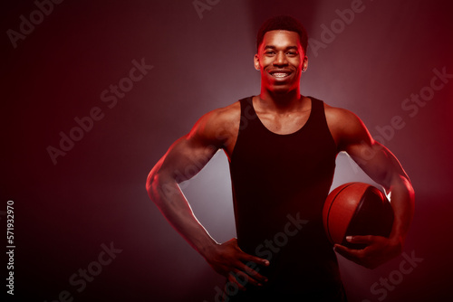 Basketball player side lit with red color holding a ball against smoke background. African american man with teeth bracelets smiling..