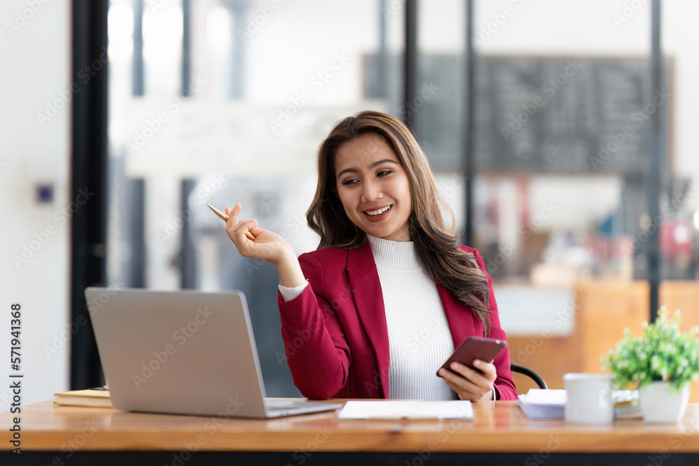 Smiling beautiful Asian businesswoman analyzing chart and graph showing changes on the market and holding smartphone at office