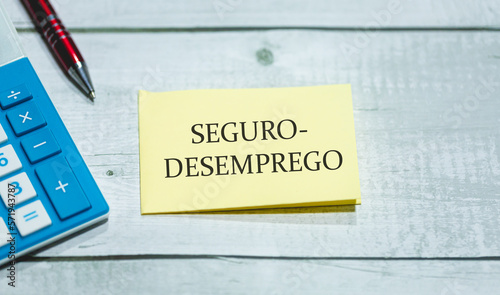 A palavra seguro-desemprego em Português do Brasil escrita em um pedaço de papel que está sobre uma mesa de madeira. Uma calculadora e uma caneta na composição. Economia brasileira. photo