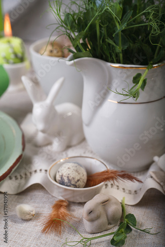 Easter table setting with fresh green plants, feathers, easter bunny and quail egg on white table.