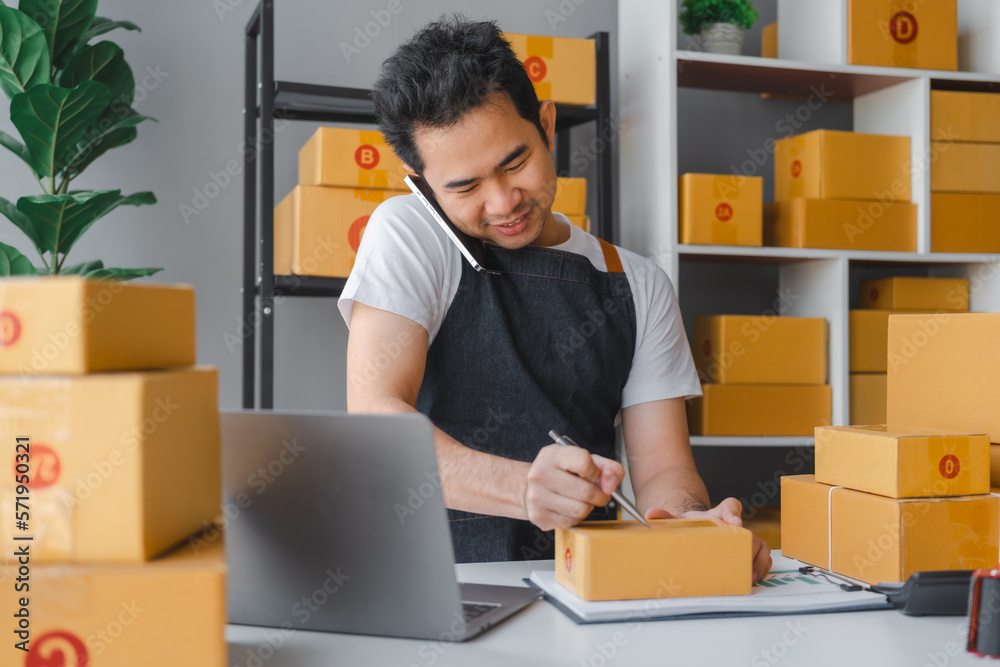 Young asian sme business man in apron working at warehouse home office freelancer retail logistic company with package box checking customer address. Young asian sme business man in apron working at warehouse home office freelancer retail logistic company with package box checking customer address.