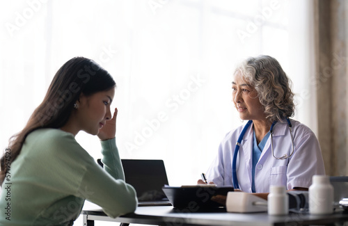 Woman senior doctor is Reading Medical History of Female Patient and Speaking with Her During Consultation in a Health Clinic. Physician in Lab Coat Sitting Behind a Laptop in Hospital Office