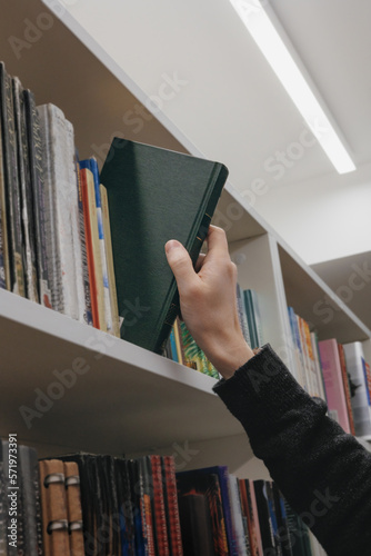 close-up of hand, young adult man stands in library in middle of shelves and takes book. reading and recreation, education on university campus, free study of exact sciences, philosophy. bookstore