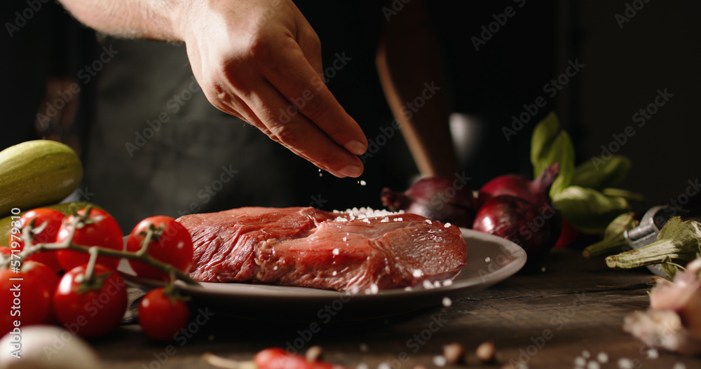 Chef applying grained salt and pepper on raw piece of steak. Cooker preparing meat on professional kitchen table with various vegetables. Close up