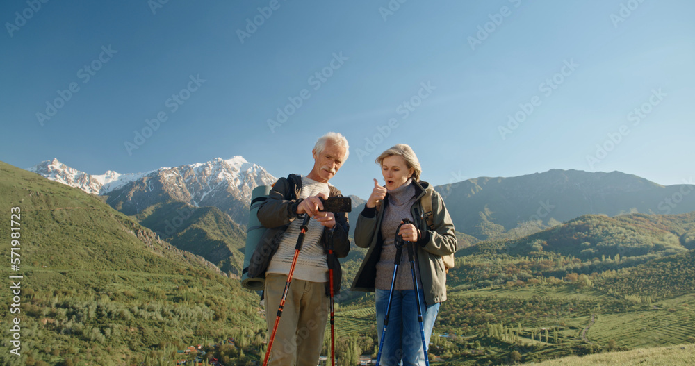 Mature caucasian couple travelling together, having a nordic walking hike in spring mountains, then stopping to take a picture on smartphone, spending time after retirement - recreational pursuit