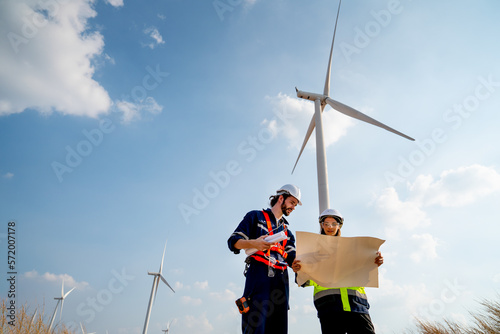 Lower view of Caucasian engineer or technician man and woman discuss together using drawing paper and stay in front of row of windmill or wind turbine with blue sky.